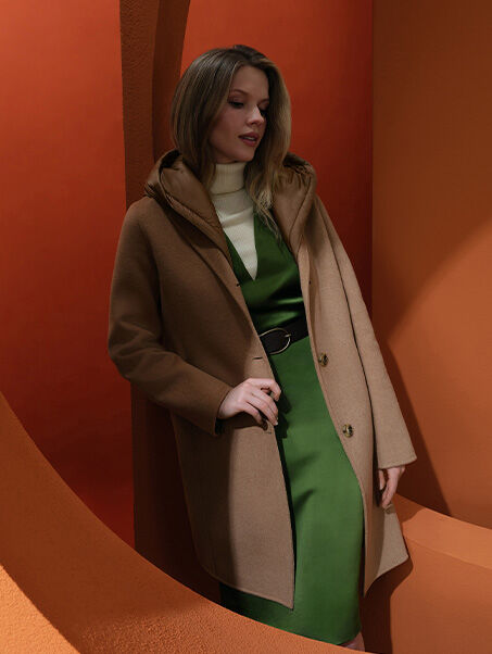 Woman standing between two orange arches with straight legs wearing a white turtleneck, green dress and camel coat with hood | GEOX | GEOX